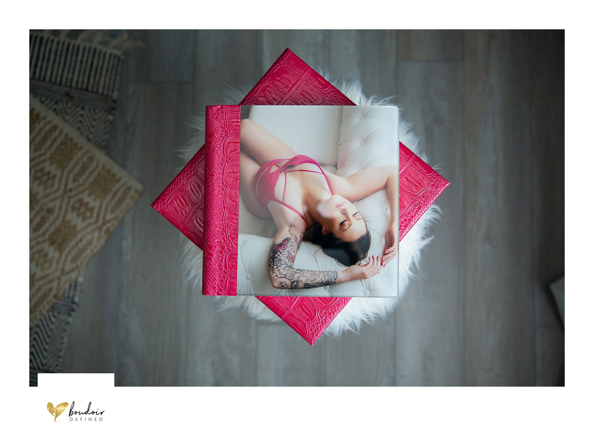 A bright pink, 8x8 boudoir album is displayed on top of a matching album case. The cover image is of a woman lying down on a white chesterfield sofa in a pink bodysuit, sporting a tattoo on her left arm.