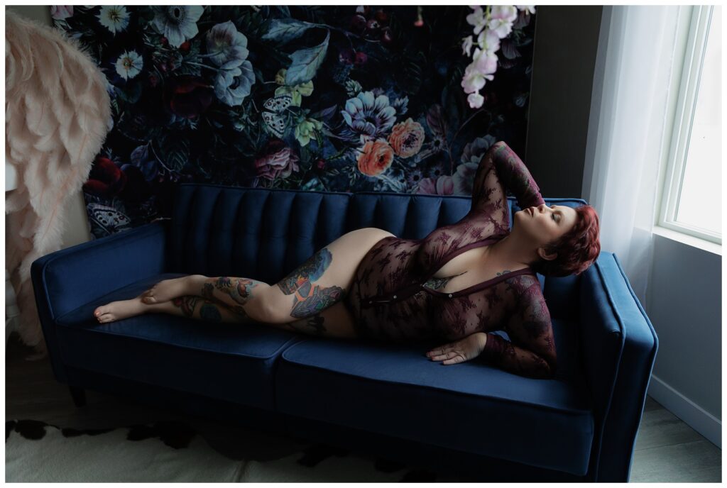Woman in sheer maroon lace bodysuit with a plunging neckline, posing on her side on a deep blue velvet couch in a softly lit room with a floral backdrop.