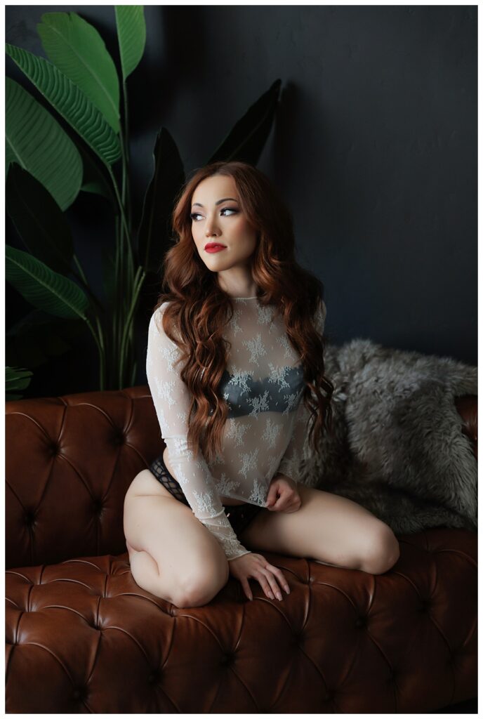 Long Haired Red-Headed woman on knees on a brown leather buttoned sofa. She's wearing a lace long sleeved top.