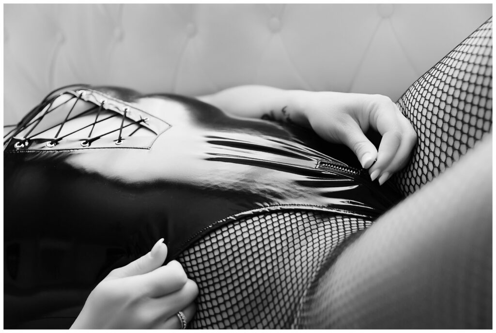 Detailed black and white Boudoir Photograph of woman unzipping bottom of pleather bodysuit in fishnet stockings.