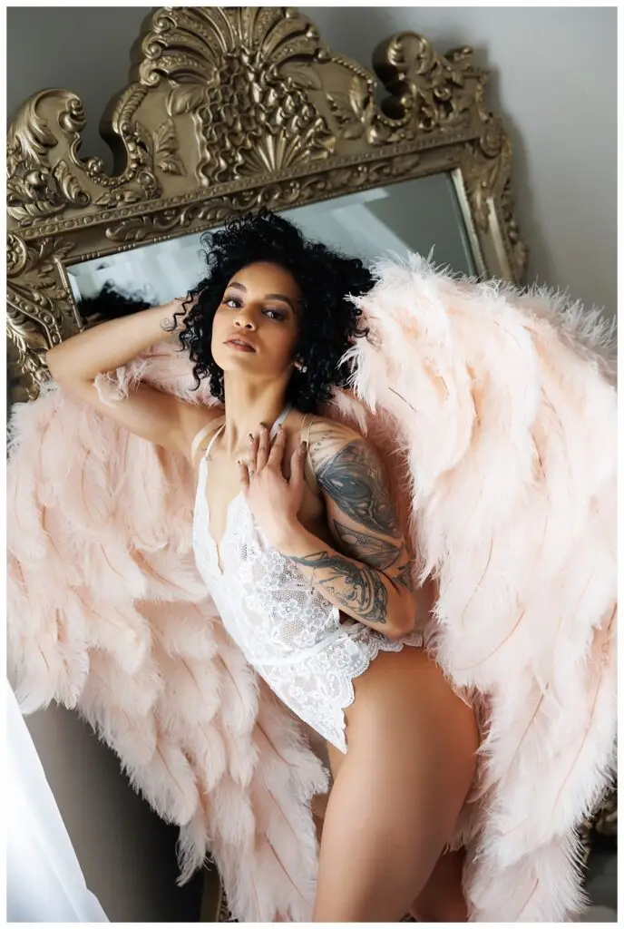 African American Woman with afro hair standing in front of mirror with blush colored wings in Boudoir Defined's Boise, Idaho Boudoir Studio.