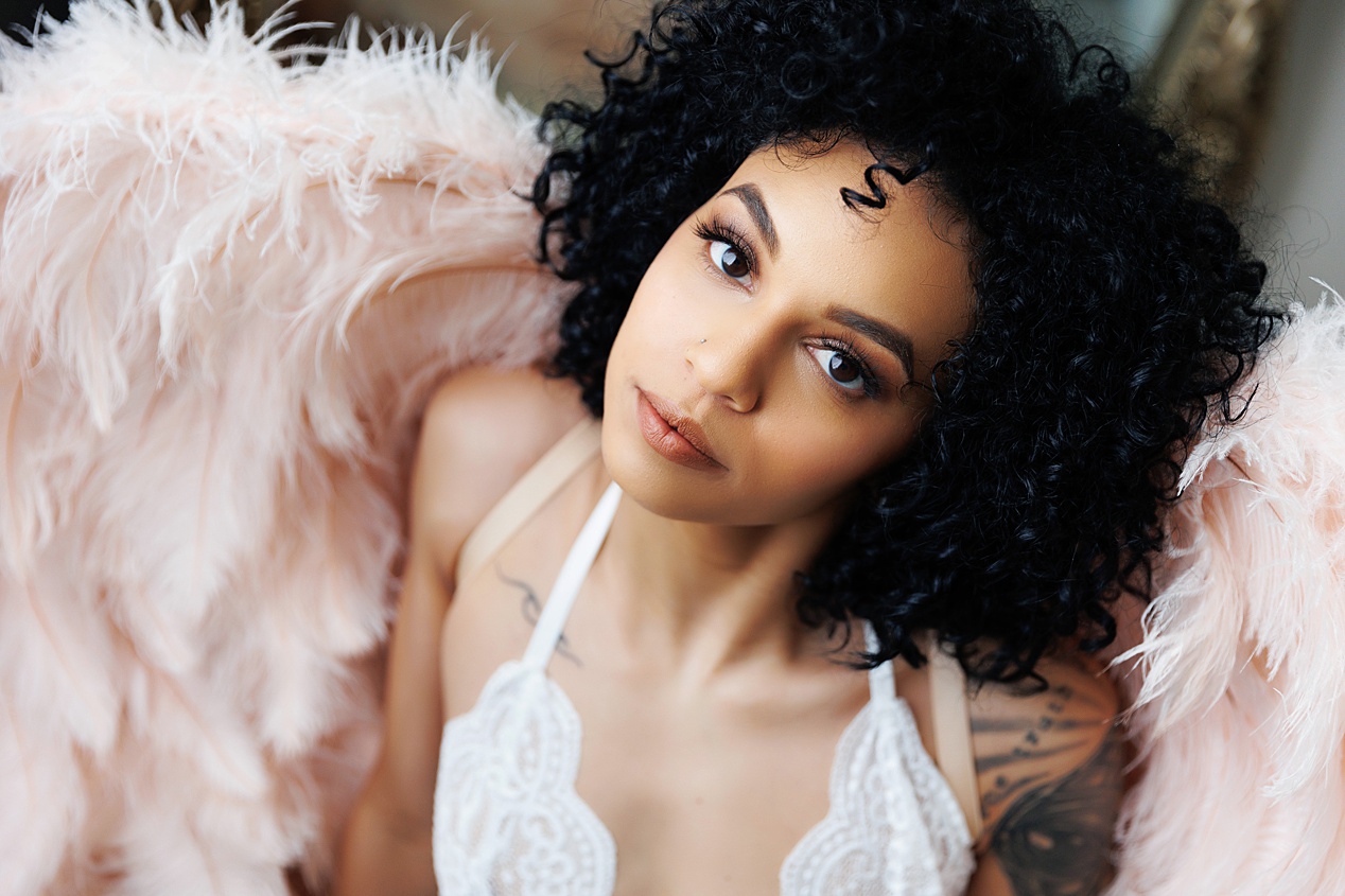 African American woman with beautiful curls looking up at the camera in pink wings at her boudoir session with Boudoir Defined in Boise Idaho